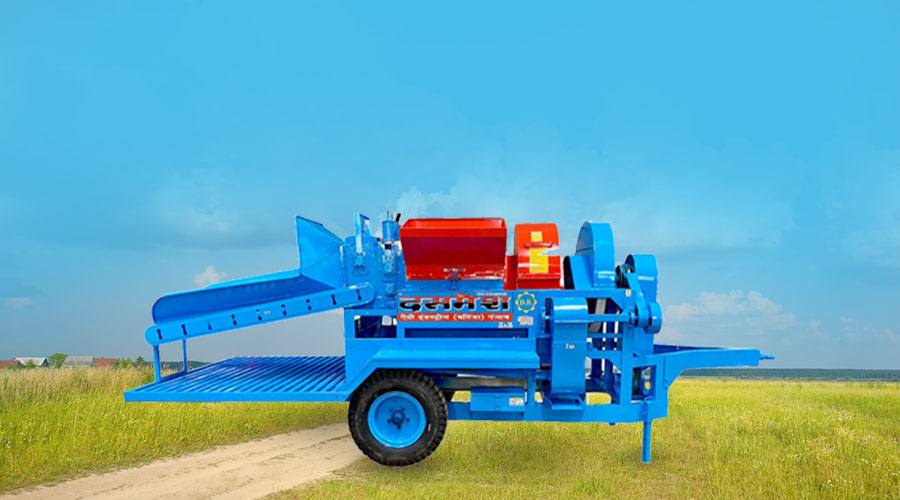 A blue agricultural thresher machine with a red motor, placed on a dirt path in a grassy field under a clear blue sky.