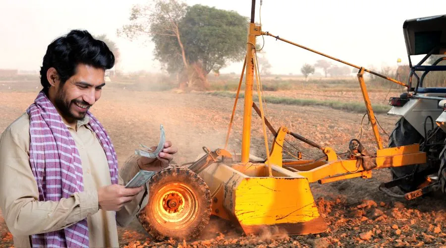 Happy Indian farmer counting money while standing near a tractor and farming equipment in a rural field.